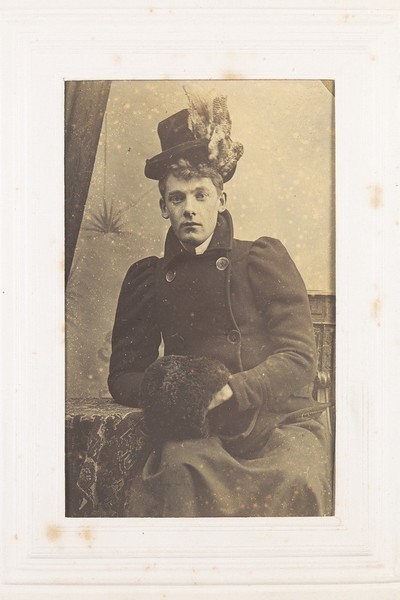 Download the full-sized image of A man in drag, seated, wearing dark clothing and a black hat. Photograph, 189-.