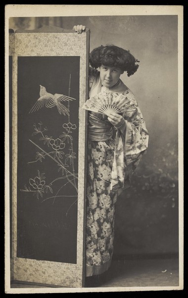 Download the full-sized image of A man in drag, wearing a kimono; posing from behind a screen. Photographic postcard, 1906.