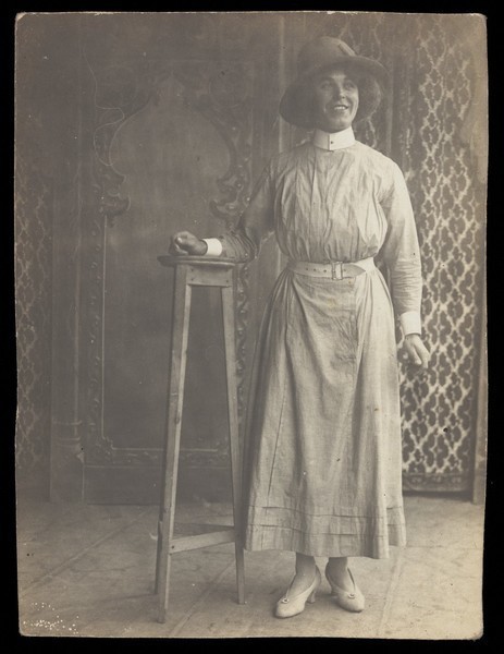Download the full-sized image of A sailor in drag leans on a high stool, in front of patterned scenery. Photographic postcard, 191-.