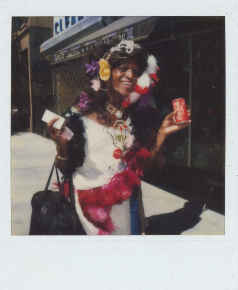 Download the full-sized image of A Photograph of Marsha P. Johnson Wearing a White Dress and Feathered Accessories, Holding a Coca-Cola Can