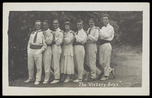 Download the full-sized image of Entertainers, one in drag, pose in a line in front of foliage. Photographic postcard, 191-.