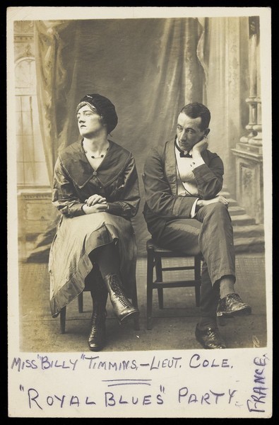 Download the full-sized image of Two soldiers, one in drag called "Miss Billy Timmins", are seated in front of a painted backdrop. Photographic postcard, 1917.