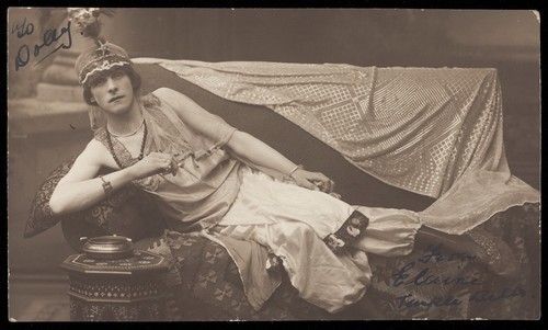 Download the full-sized image of A man in drag poses wearing delicate attire; curled up on a piece of furniture. Photographic postcard, 191-.
