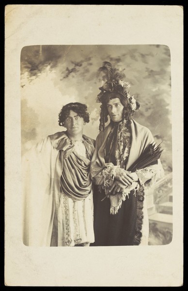 Download the full-sized image of Two men in drag, one wearing a detailed feathered head garment, pose for a portrait. Photographic postcard by F. Wood, 190-.