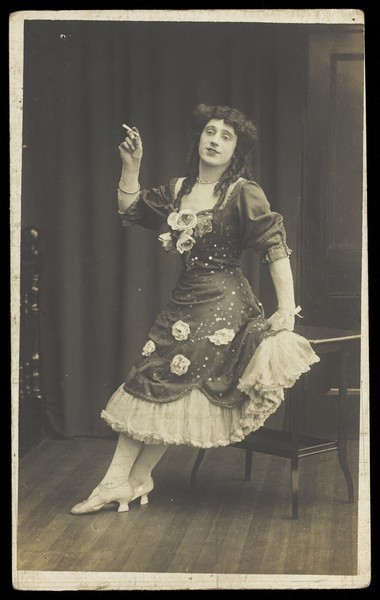 Download the full-sized image of A performer in drag, wearing a large flowery dress, poses with a cigarette on stage. Photographic postcard, ca. 1908-1910.