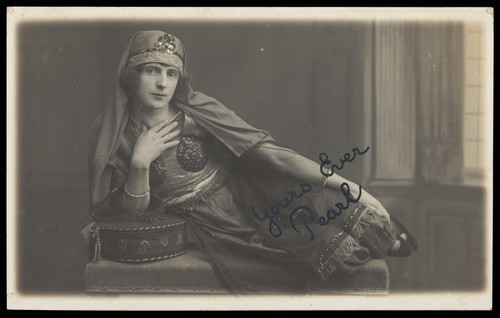 Download the full-sized image of A man in drag, poses wearing delicate attire; curled up on a piece of furniture. Photographic postcard, 191-.