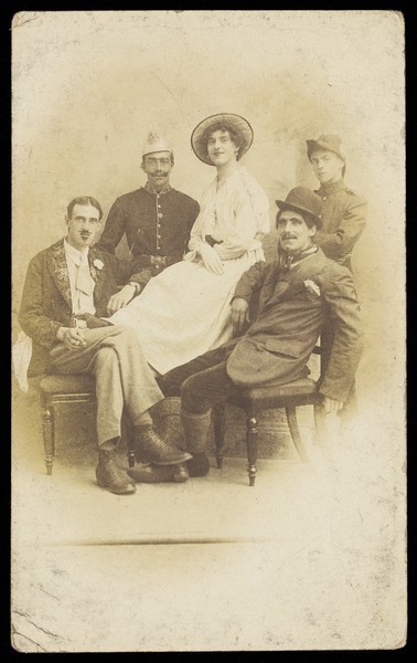 Download the full-sized image of Amateur actors sitting down, surrounding a man in drag seated in the centre. Photographic postcard, 191-.