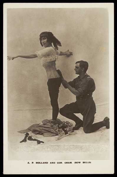 Download the full-sized image of Con Oram laces A.P. Holland (in drag) into a boned corset in a show for the Bow bells concert party. Photographic postcard, 191-.