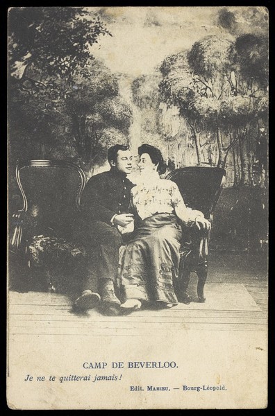 Download the full-sized image of Two soldiers, one in drag, sit close to each other on stage, in front of a detailed backdrop of trees at Beverlo Camp, Belgium. Process print, 190-.