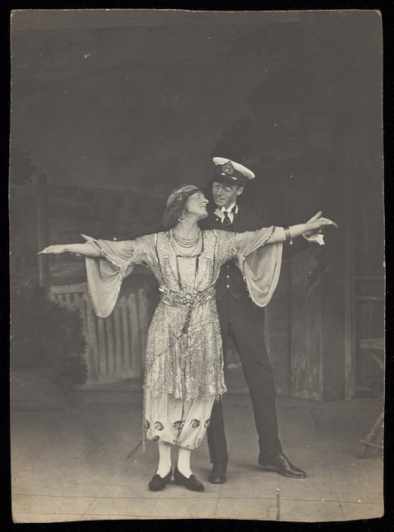 Download the full-sized image of A couple, one in drag, pose together on stage. Photographic postcard, 191-.
