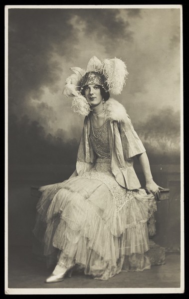 Download the full-sized image of A young man in elaborate drag sits in front of a painted sky backdrop. Photographic postcard, 1934.