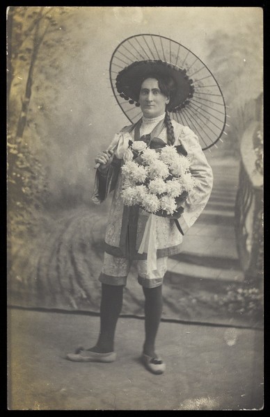 Download the full-sized image of An amateur actor in drag, holding flowers and a parasol, poses in front of delicately painted backdrop. Photographic postcard, 191-.