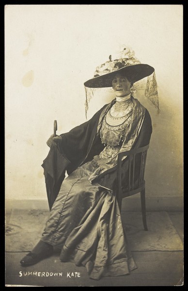 Download the full-sized image of A convalescent soldier (Lance-Corporal R.C. Thomas) poses in drag, wearing a silk dress and sitting with a mourning veil. Photographic postcard, 1916.